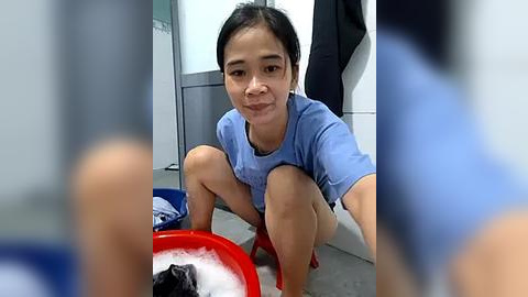 Media: Video of a young woman with short black hair and medium skin tone, squatting in a bathroom, wearing a blue t-shirt and shorts, washing a black dog in a red plastic basin.