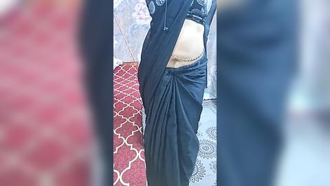 Media: Video of a person in a black, pleated saree standing on a red carpet with white geometric patterns, partially obscured by another person in the background.