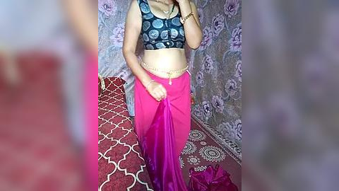 Media: Video of a woman in a blue patterned crop top and bright pink saree with a gold belt, standing in a room with floral-patterned purple curtains and a red-patterned carpet.