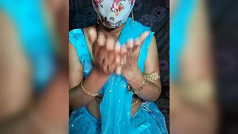 Media: A video of a woman in a blue saree with gold accessories, wearing a floral face mask, hands clasped together, blurred background.