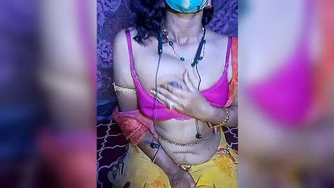 Media: Video of a woman with medium brown skin, wearing a pink bra, yellow sari, and blue mask, sitting on a patterned floor. She covers her breasts with her hand.