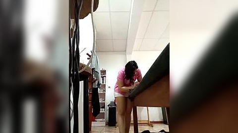 Media: A video shows a woman with dark hair, wearing a pink shirt and shorts, bending over a desk in a brightly lit, modern office with white walls and ceiling tiles.