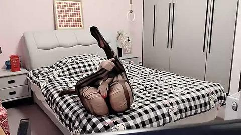 Media: Video of a woman in black stockings and a brown skirt, lying on a black-and-white checkered bed in a modern, minimalist bedroom with white furniture and pink walls.