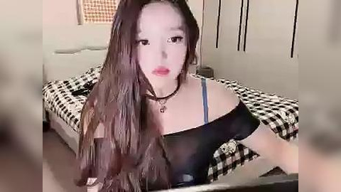 Media: Video of a young East Asian woman with long, wavy brown hair, wearing a black off-shoulder top and a black choker, sitting on a bed with black-and-white checkered bedding, in a dimly lit room with beige walls.