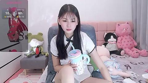 Media: Video of an East Asian woman with straight black hair, wearing a white short-sleeved shirt, drinking from a blue and white bubble tea cup, in a pink-tufted bed, surrounded by plush toys, including a pink polka-dotted bear.