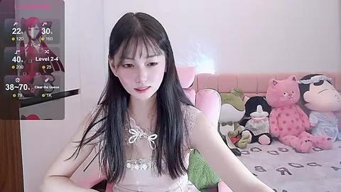 Media: A video of an East Asian woman with long black hair, wearing a white lace dress, sitting on a pink gaming chair in a bedroom with plush toys and a TV displaying game statistics.