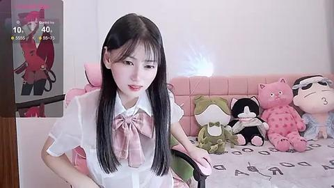 Media: Video of a young Asian woman with long black hair and fair skin, wearing a white blouse and pink bow, sitting in a pink cushioned chair. Background features stuffed animals and a TV screen displaying an anime character.