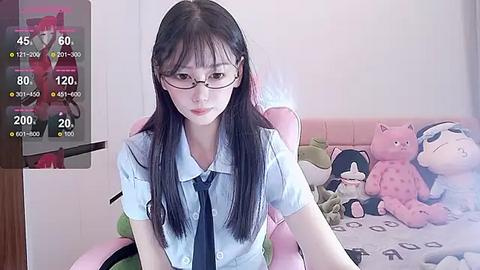 Media: Video of an Asian woman with long black hair and glasses, wearing a light blue shirt and tie, seated in a pink gaming chair, surrounded by plush toys and a TV displaying a character.