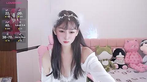 Media: Video of an East Asian woman with long black hair, wearing a white off-shoulder dress, sitting on a pink bed with stuffed animals.