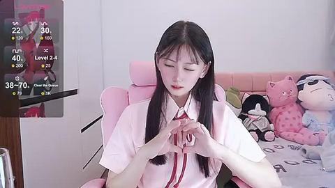 Media: Video of an Asian woman with long black hair, wearing a pink school uniform, seated on a pink gaming chair. She's playing a game on a monitor displaying a character in a red uniform. Background includes plush toys and a desk.