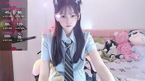 Media: A video of a young, slender Asian woman with long, straight black hair, wearing a light blue short-sleeve shirt, black tie, and black headband, sitting on a bed surrounded by plush toys, captured on a smartphone.