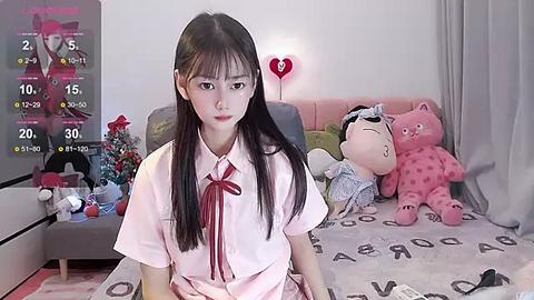 Media: Video of an East Asian woman with long black hair and bangs, wearing a pink school uniform, sitting on a gray couch with plush toys. Background includes a wall calendar and a heart-shaped light.