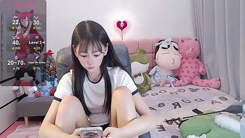 Media: Video of an Asian woman with long black hair, wearing a white t-shirt, sitting on a bed with plush toys, playing a smartphone game.