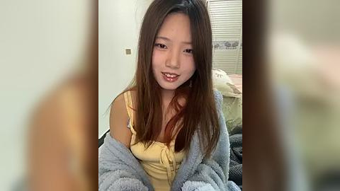 Media: Video of an Asian woman with long, straight, brown hair and fair skin, wearing a yellow top and grey cardigan, smiling softly. Background features a blurred bedroom with white walls and a bed.