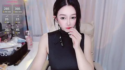 Media: A video of a pale-skinned East Asian woman with long black hair, wearing a black sleeveless top, sitting in a modern room with a vanity and makeup items.