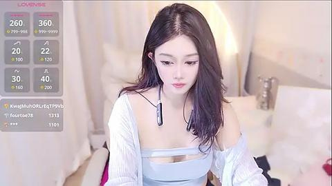 Media: A video of a young Asian woman with long, straight black hair, fair skin, and a slender physique, wearing a light blue off-shoulder top, sitting on a white bed in a softly lit bedroom.