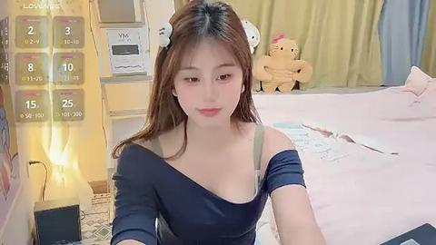 Media: Video of an East Asian woman with light skin, long brown hair, and bunny ears, wearing a black off-shoulder top, sitting on a pink bed with teddy bears, in a cozy room.