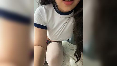 Media: Video of a young woman with long dark hair, wearing a white T-shirt with blue trim, squatting on a bed, partially obscured by a blurry object.