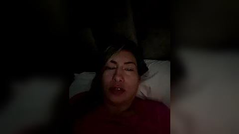 Media: Video of an Asian woman with long black hair, wearing a red shirt, lying on a white pillow, eyes closed, dimly lit room, blurred foreground.