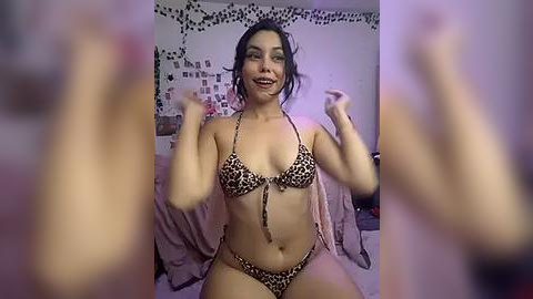 Media: Video of a smiling, dark-haired woman in leopard-print bikini, posing with fists raised in a playful manner in a bedroom with pastel walls and a bed.