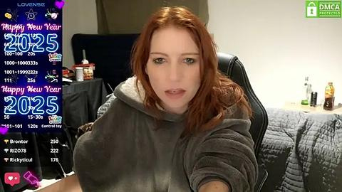 Media: A video of a fair-skinned woman with red hair in a gray hoodie, sitting on a black chair in a cozy room. The background includes a bed, a desk with a monitor, and various personal items.