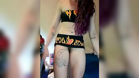 Media: Video of a pale-skinned woman with long, wavy purple hair, wearing a black and yellow flame-patterned bikini, showcasing tattoos on her thighs and arms. Background includes a blurred person in red, and a dark blue surface.