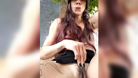 Media: A video of a woman with long, dark hair, tattoos, and glasses, sitting outside, partially undressed, exposing her pubic hair.