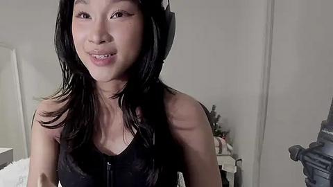 Media: Video of an Asian woman with long black hair, wearing a black top, smiling. She is indoors, in a dimly lit room with a white wall and a camera visible.