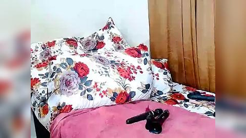 Media: A video of a neatly made bed with a white quilt featuring red and purple floral patterns, surrounded by soft, floral-patterned pillows, with a pink blanket at the foot. A black handgun lies on the blanket.