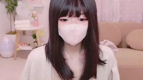 Media: Video of an Asian woman with long black hair, wearing a white face mask and robe, in a pastel-colored, cozy room with a beige sofa and potted plants.