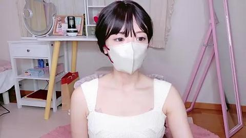 Media: Video of an East Asian woman with short black hair, wearing a white mask and white lace top, in a cluttered bedroom with a white desk, fan, and pink stand.