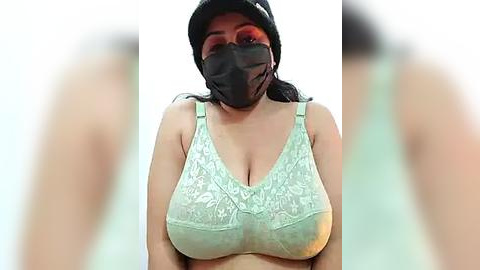 Media: Video of a South Asian woman with medium skin tone, wearing a black face mask, black hat, and a light green lace bra, showcasing medium-sized breasts. Background is blurred.