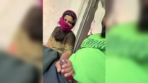 Media: Video of a woman in a pink headscarf and beige shirt, holding a baby\u2019s penis, with a blurred background and green shirt in foreground.