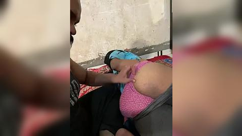 Media: A video of a woman breastfeeding her baby in a dimly lit room with a beige wall and red-patterned fabric. The woman wears a pink polka-dot top and blue headscarf.