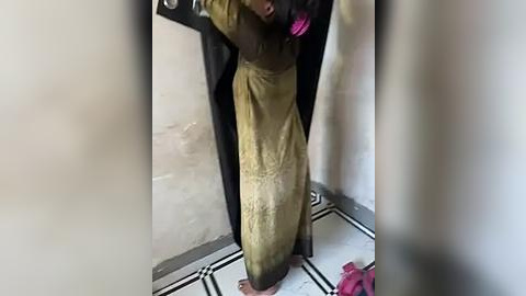 Media: A video of a woman in a beige, floral-patterned dress with a black, long-sleeved overcoat, standing barefoot on a tiled floor in a dimly lit, narrow corridor.