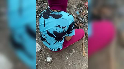 Media: Video of a person wearing a blue and black floral-patterned shirt and magenta pants, sitting on a dirt ground with scattered debris.