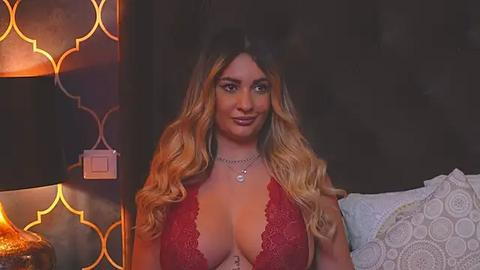 Media: Video of a light-skinned woman with long, wavy blonde hair, wearing a red lace bra, smiling in a dimly lit bedroom with warm, geometric-patterned wallpaper.