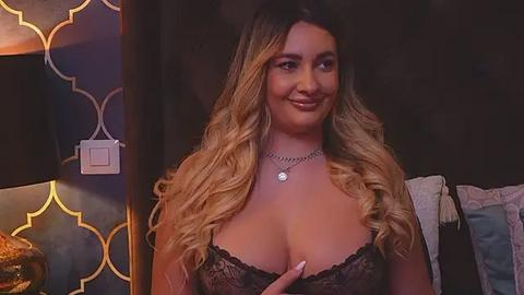 Media: Video of a smiling, light-skinned woman with long, wavy blonde hair, wearing a black lace bra that reveals ample cleavage. She's in a dimly-lit room with geometric-patterned wallpaper and a white light switch.