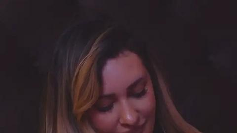 Media: Video of a young woman with long, wavy hair featuring blonde highlights, closed eyes, and a soft smile, set against a dark, blurred background.