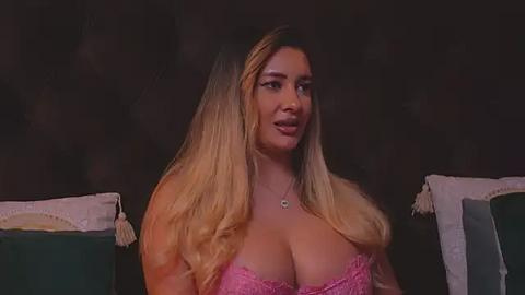 Media: Video of a curvy, light-skinned woman with long, blonde hair, wearing a pink lace bra, sitting against a dark, textured wall with green and beige pillows.