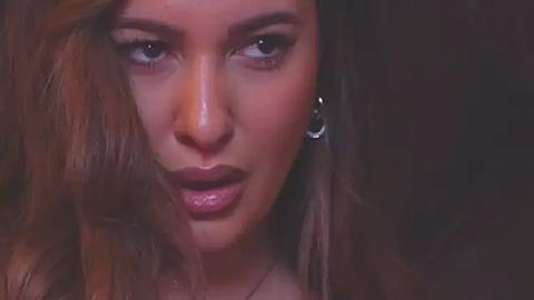 Media: Video of a close-up of a woman with long, wavy brown hair and light skin, wearing a silver hoop earring, looking serious with slightly parted lips. Dark background.