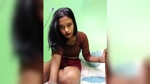 Media: A video of a young South Asian girl with straight black hair, brown eyes, and medium skin tone, wearing a maroon top and skirt, sitting on a white bed with a green backdrop.