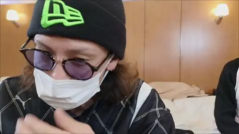 Media: Video of a woman in a black beanie, black-framed sunglasses, and a white face mask, wearing a black plaid shirt, sitting on a bed with beige walls and white sheets in a bedroom.