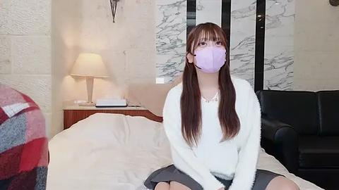 Media: Video of a young Asian woman with long brown hair, wearing a purple face mask, white sweater, and black skirt, sitting on a bed with a beige blanket, in a modern, minimalistic room with marble walls and a black leather couch.