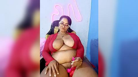 Media: Video of a plus-sized Black woman with glasses, wearing a red bikini top and panties, sitting on a chair in a room with pink neon lights and a blue wall.