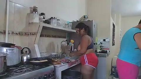 Media: Video of a woman in a kitchen, wearing a black sports bra and red shorts, cooking on a stove, with a stainless steel sink and cabinets in the background.