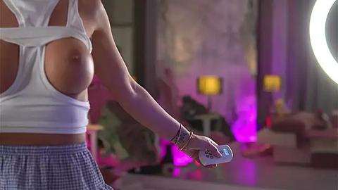 Media: Video of a woman with a white crop top revealing her left breast, holding a remote, in a dimly lit, modern living room with purple lights and a round mirror.