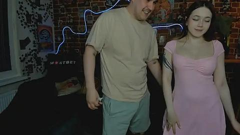 Media: Video of a smiling, slender Caucasian woman in a pink dress and a chubby, light-skinned man in a beige t-shirt and light blue shorts, both holding hands. Background includes a brick wall, blue neon lights, and a Star Trek poster.