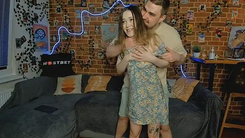 Media: Video of a joyful couple dancing in a cozy, eclectic living room with a brick wall backdrop, colorful posters, and a blue neon light.