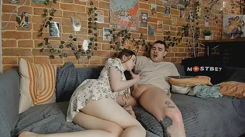 Media: Video of a young woman with light skin and brown hair kneeling on a gray couch, performing oral sex on a man with a tattooed leg, in a cozy room with brick walls, posters, and a \"MOST BET\" sign.